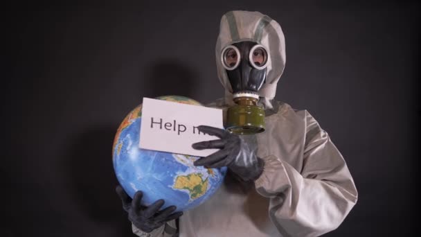 Людина в костюмі хімічного захисту і протигаз. Хлопець тримає модель Землі, глобус. Плакат з наступним написом прикріплений до земної кулі: HELP ME. — стокове відео