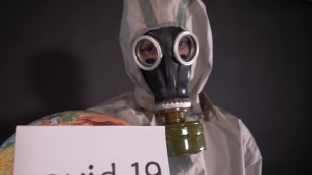 Man in a chemical protection suit and a gas mask. The guy is holding a model of the Earth, a globe. A placard with the following inscription is attached to the globe: covid-19. — Stock Video