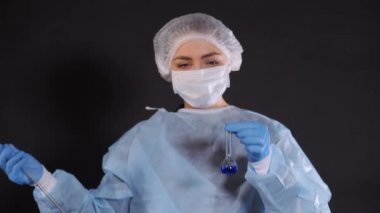 Bir damla mavi sıvı, ısıya dayanıklı su ölçme cihazının cam tüpünden şeffaf bir kaba düşer. Koruyucu giysili genç bir kadın. Koruyucu bir yüzün üzerinde