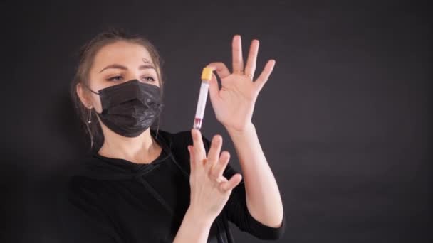 Vrouw met een zwart beschermend masker met de woorden "kovid-19" op haar voorhoofd geschreven. Het meisje houdt een reageerbuis vast met biomateriaal.. — Stockvideo