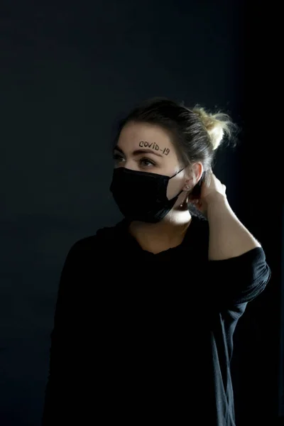 Femme dans un masque de protection noir avec les mots covid-19 écrit sur son front. Fille pousse une mèche de cheveux de son visage, exposant ainsi le front avec l'inscription . Images De Stock Libres De Droits