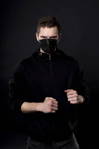 Muž v černém v bojové póze se zaťatými pěstmi. Na obličeji mladého muže mu černá lékařská maska zakrývá nos a ústa.. Stock Snímky