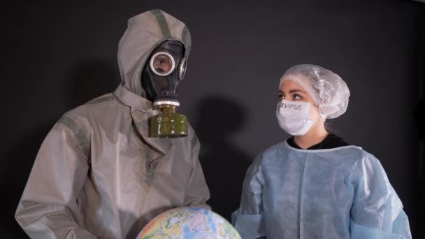 化学防護服の男とガスマスクだ。男は地球、地球のモデルを保持している。保護服を着た若い女性。保護医療マスクの顔に。治療法 — ストック動画