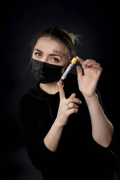 額にkovid-19と書かれた単語と黒の保護マスクの女性。女の子が持っています試験管とともに生体材料. — ストック写真