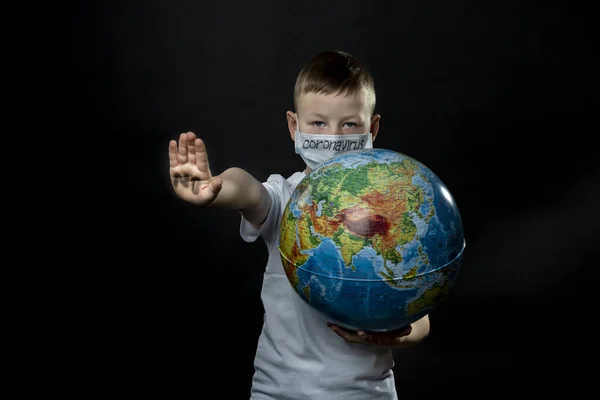 Baby reaches forward hand STOP, NO. Boy is holding a globe, a model of the planet Earth. Medical mask on the child\'s face. On the white mask is a black inscription Coronavirus. Boy is careful of the virus. Pandemic March 2020. Coronavirus. COVID-19