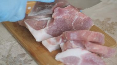 Yemek yapmak, taze et kesmek. Eldiven giymiş eller ve tek kullanımlık mavi bir bornoz. Tahtada domuz eti dilimlemek. Bıçakla parçalara ayırmak..