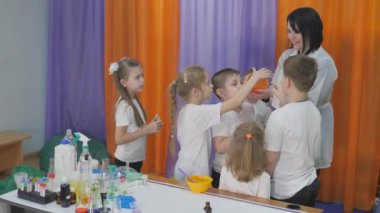 Çocuklar için kimyasal deneyler. İnsanlar elastik kabarcıkları şişirir. Çocukların parlak duyguları. Çocuklar için eğlenceli deneyler. Bir kadın bilişsel bilim dersleri veriyor. Çocuklar için eğlenceli oyunlar. Çocuklar ellerinde kimya tabaklarıyla masada dururlar..