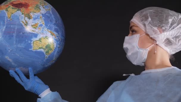 穿着浅色防护服的女孩。人的脸上有医疗面具.她的头发上戴着一顶医疗帽.一个年轻的女人拿着一个地球模型. — 图库视频影像