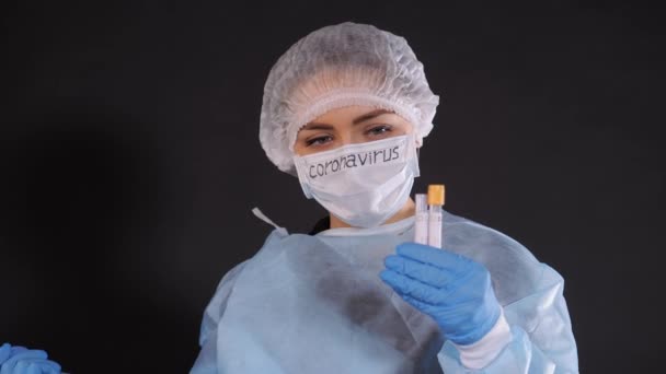 Το κορίτσι εξετάζει ένα δοκιμαστικό σωλήνα με βιοϋλικό υλικό. Νεαρή γυναίκα με προστατευτική ενδυμασία. Στο πρόσωπο μιας προστατευτικής ιατρικής μάσκας. Μια σταγόνα υγρού πέφτει από το σωλήνα μιας γυάλινης θερμοανθεκτικής μέτρησης νερού — Αρχείο Βίντεο