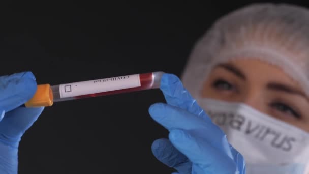 Girl examina un tubo de ensayo con biomaterial. Una joven con ropa protectora. En la cara de una máscara médica protectora. Las manos están vestidas con guantes de látex azul. En la máscara blanca-negro — Vídeos de Stock