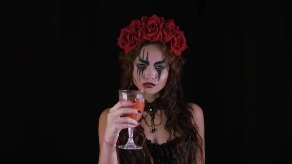 Maquillaje fácil de Halloween. La chica con la foto en la cara. La novia de los demonios con una corona de flores rojas en la cabeza. Mujer bebe de un vaso de bebida roja ofreciendo a beber para el espectador . —  Fotos de Stock