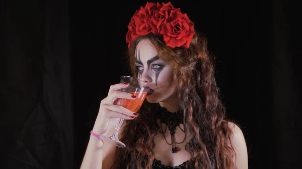 Lehký Halloweenský makeup. Dívka s obrázkem na tváři. Ďábelská nevěsta s věnečkem červených květin na hlavě. Žena pije ze sklenice červeného nápoje a nabízí divákovi pití. — Stock fotografie