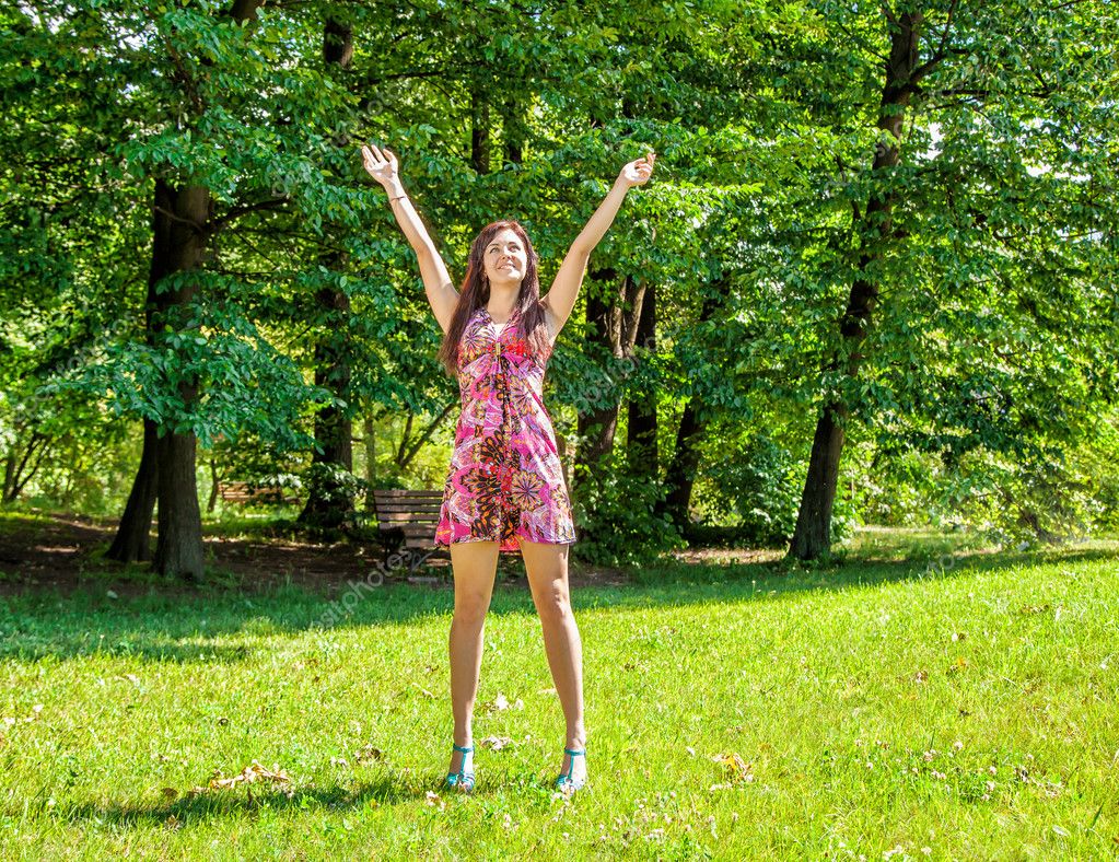 Young beautiful girl in red dress standing hands up in the park on summer d...