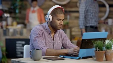 4k genç rahat kulaklık üzerinden müzik ve kafede dizüstü bilgisayarda çalışan adam