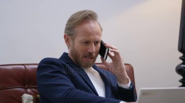 Çekici akıllıca giyimli adam evde telefonda konuşurken ve dizüstü bilgisayarda çalışma