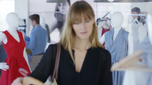 ファッション衣料品店でミラーにして衣服を保持している女性客 ショッピング バック グラウンドで他のお客様と鏡の視点から見た — ストック動画