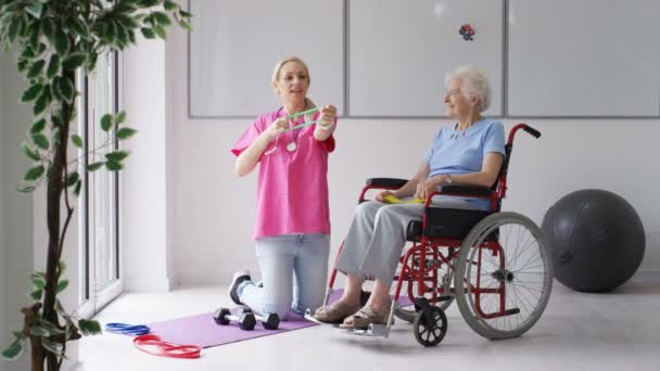 有料老人ホームで看護師は車椅子で年配の女性を示す抵抗バンドといくつかの演習 — ストック動画