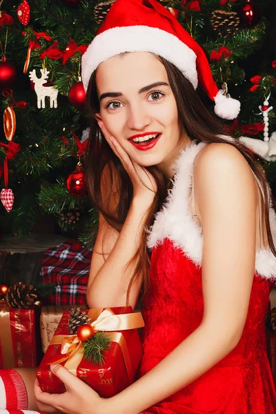 Young happy woman in red santa hat and red dress sitting near ne — Stock Photo, Image