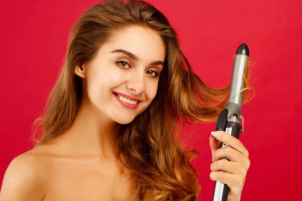 Молодая улыбчивая женщина делает кудрявые волосы с помощью уловки на красном фоне — стоковое фото