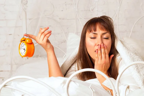 Young attractive sleepy woman laying in bed and holding clock in — Stock Photo, Image