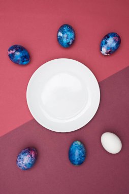 Paskalya bayramı renkli yumurtalar ve mor arka planda beyaz tabak. Üst görünüm.