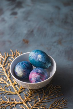 Beyaz bir kasede Paskalya yumurtaları ve mavi desenli bir yüzeyde altın bir dal..