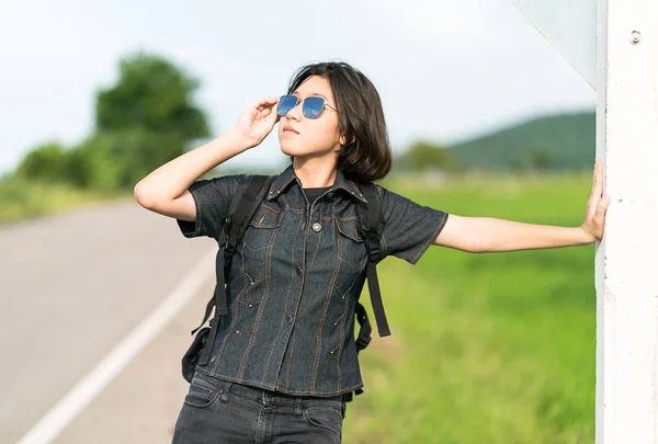 Mujer de pie con mochila autostop a lo largo de un camino —  Fotos de Stock