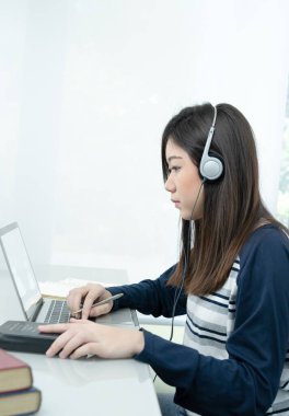 Genç bir kadın oturma odasında oturuyor ve kulaklık takıyor. İnternette öğreniyor. Masada dizüstü bilgisayar kullanıyor.