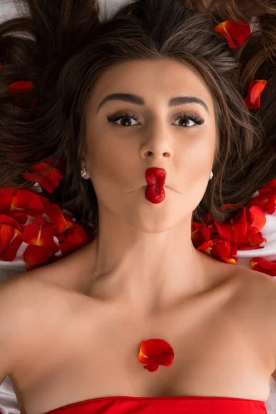 Bruna in rosso con labbra emotive sul letto con petali — Foto Stock