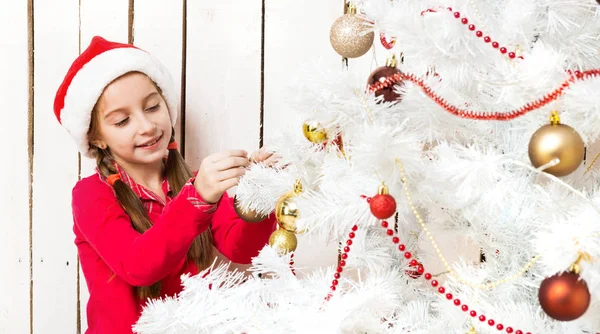 Маленькая девочка в красной шляпе Санты, украшающая новогоднюю елку — стоковое фото
