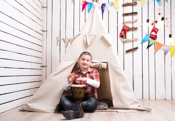 Niño sentado en el suelo y tocando el tambor cerca de wigwam — Foto de Stock