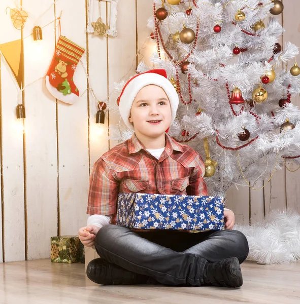 Bambino in cappello rosso che prende la scatola regalo vicino al nuovo albero di anno — Foto Stock