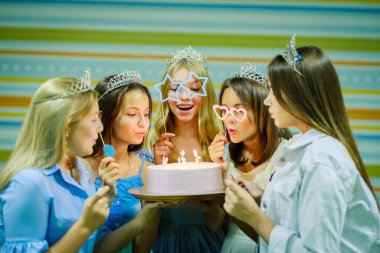 Güzel, gülümseyen, elbiseli ve taçlı genç kızlar pastayı bir arada tutuyor ve doğum günü partisinde mumları üflüyorlar.