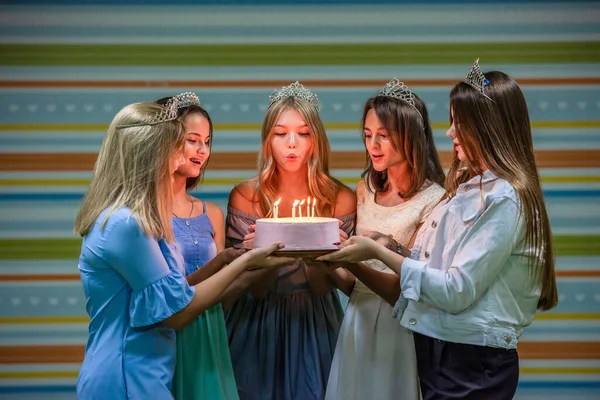 Ganska leende tonårstjejer i klänningar och kronor håller ihop tårtan och blåser ljusen på födelsedagsfesten — Stockfoto