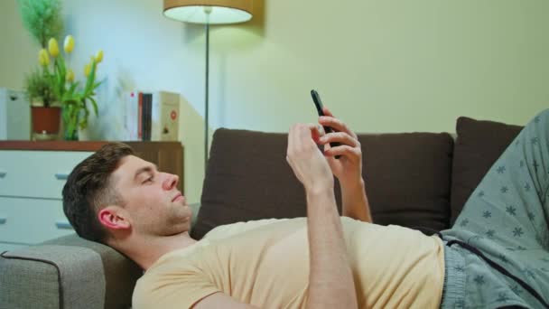 Hombre acostado en el sofá y el uso de teléfono móvil — Vídeos de Stock