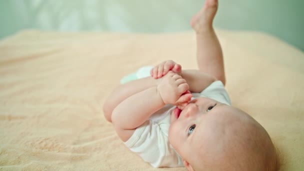 Bebé acostado en la cama en casa comiendo su pie — Vídeos de Stock