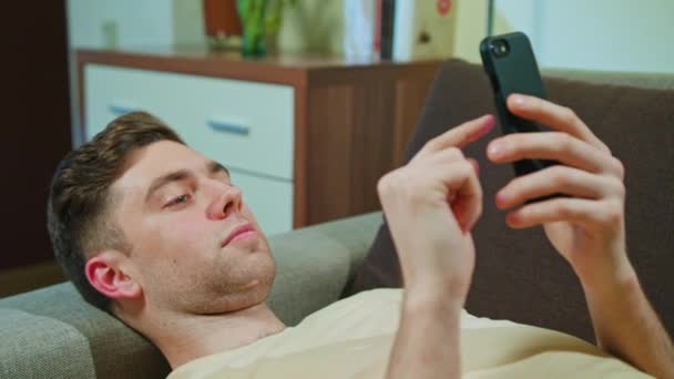 Человек лежит на диване и пользуется мобильным телефоном — стоковое видео