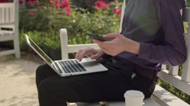 Genç adam parkta bir dizüstü bilgisayar ve bir telefon kullanma