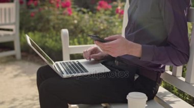 Genç adam parkta bir dizüstü bilgisayar ve bir telefon kullanma