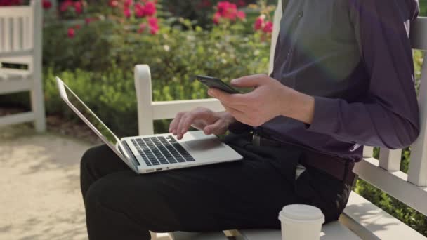 Hombre joven en el parque usando un ordenador portátil y un teléfono — Vídeo de stock