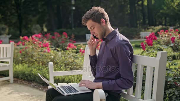 Νεαρός άνδρας στο πάρκο χρησιμοποιώντας ένα φορητό υπολογιστή και ένα τηλέφωνο — Αρχείο Βίντεο