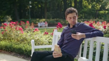 Genç adam parkta bir telefonu kullanarak ve yeme
