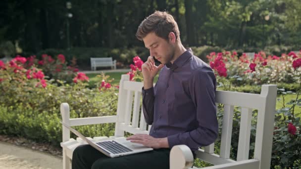 Jeune homme dans le parc utilisant un ordinateur portable et un téléphone — Video