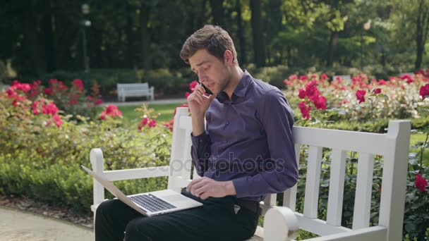 Молода людина в парку використовуючи ноутбук і телефон — стокове відео