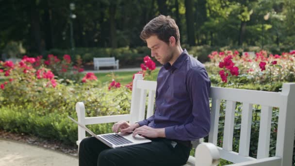 Yorgun genç adam parkta bir dizüstü bilgisayar kullanarak — Stok video