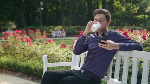 Joven en el parque usando un teléfono y bebiendo — Vídeos de Stock