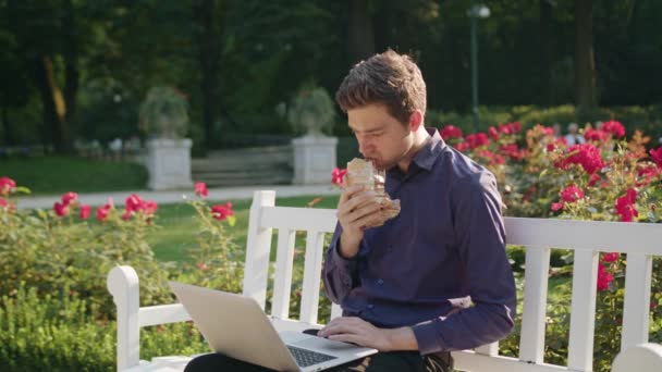 Młody mężczyzna w parku za pomocą laptopa i jedzenie — Wideo stockowe