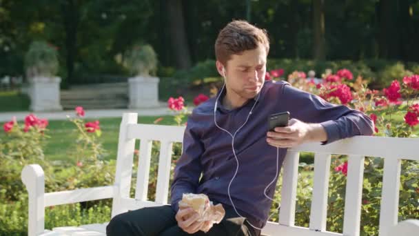 Joven en el parque escuchando música y comiendo — Vídeos de Stock