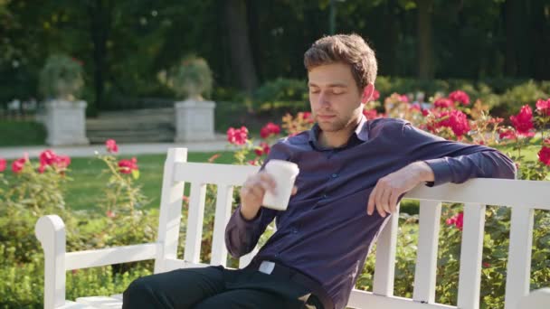 Молодой человек пьет кофе в парке — стоковое видео