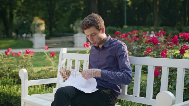 Молодой человек перекусывает в парке и читает — стоковое видео